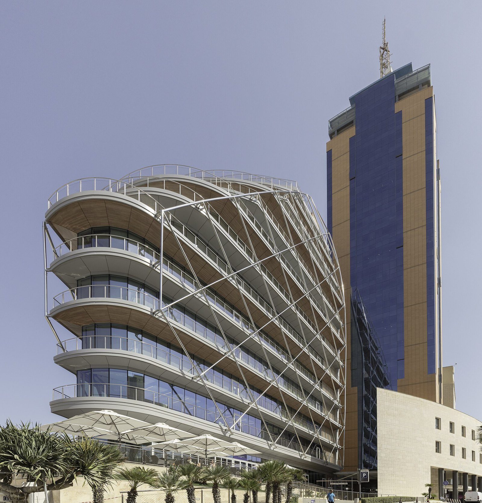 Edificio curvo de balcones y estructura metálica junto a una torre azul y dorada, con palmeras en primer plano, en St. Julian's, Malta