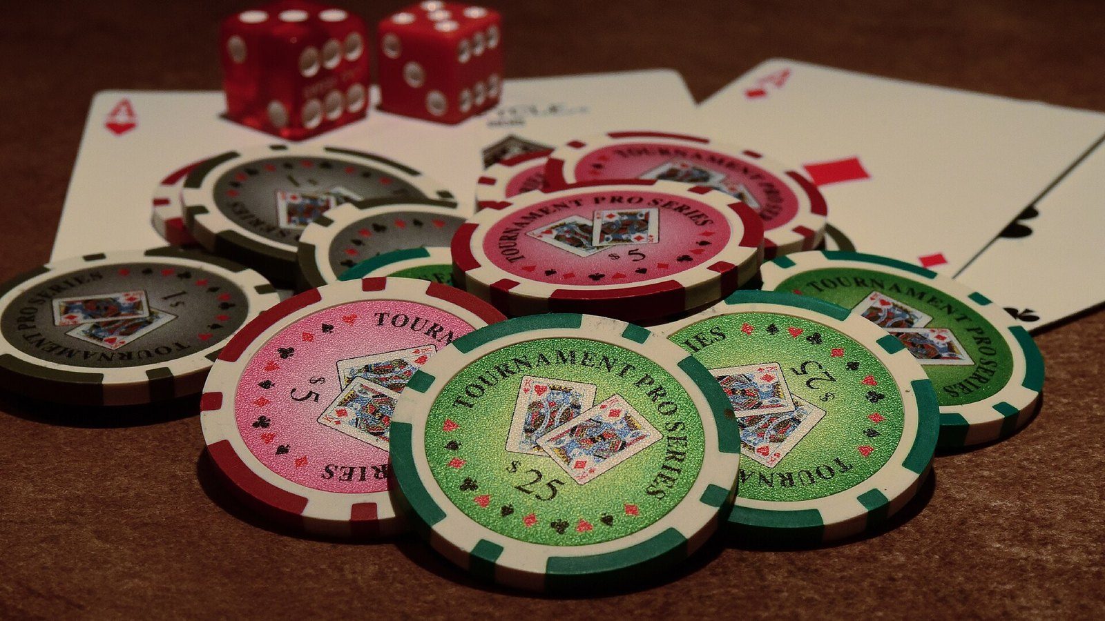 Fichas de póker verdes, rosas y rojas apiladas sobre cartas y dos dados rojos, en una mesa de fieltro marrón
