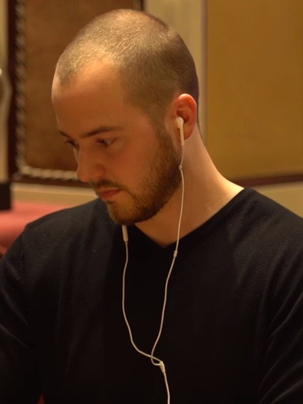 Sean Winter de perfil, con barba corta, auriculares blancos y suéter negro, mirando hacia abajo en la mesa de un torneo